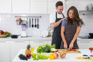 Kocası ona mutfakta sarılırken, kahvaltıda organik sağlıklı salata pişirmek için sebze doğrayan beyaz bir kadın. çift kavramı