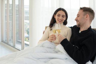 Gülümseyen yüzlü, yatakta yan yana oturan ve evde şarap içen beyaz bir çift. birliktelik konsepti