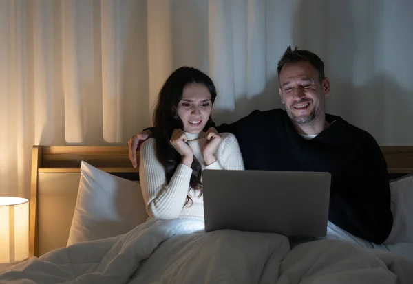 cuacasian couple watcing scary movies from streming program in computer together on bed at home in the night