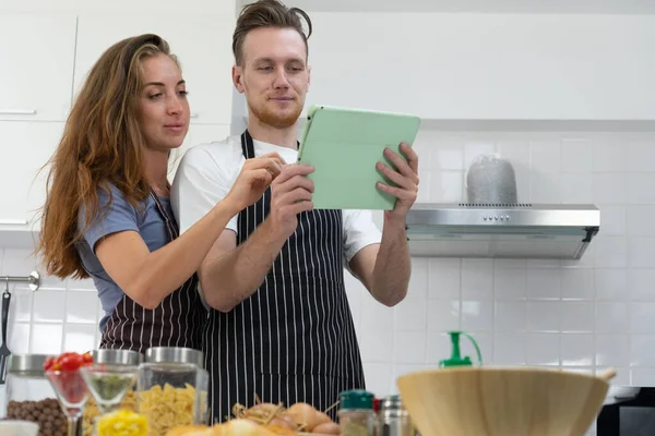 若い白人夫婦は台所で料理をする前にタブレットで新しい料理の指示を読む — ストック写真