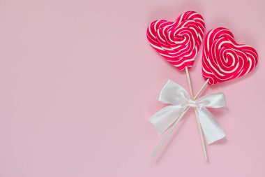 Pembe arka planda iki parlak lolipop var. Sağ tarafında beyaz bir fiyonk olan tahta bir çubuğun üzerinde kalbi olan iki parlak lolipop. Tebrik kartı. Boşluk. Üst Manzara. Düz konum düzeni. Minimalizm.
