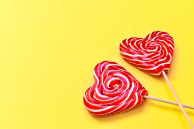 Sarı arka planda, sağ tarafında beyaz bir fiyonk olan tahta sopanın üzerinde kalbi olan iki parlak lolipop. Tebrik kartı. Yukarıdan manzara. Düz plan. Minimalizm.