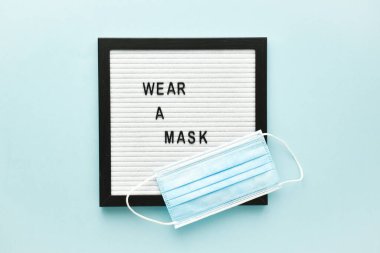 Maskeye açık mavi bir arka planda tıbbi maske ve keçeli tahta üzerine kısa mesaj konur..