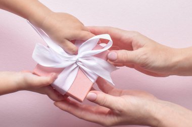 Bir çocuğun elinin en üst görüntüsü ve bir kadının eli pembe arka planda beyaz kurdeleli pembe bir hediye kutusunu geçiyor. Mutlu yıllar, mutlu anneler günü..