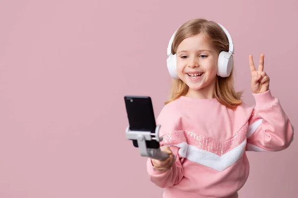 Mała dziewczynka bloger influencer nagrywa filmy z bloga na smartfonie, komunikuje się z abonentami, robi selfie, słucha muzyki ze słuchawkami, i zdalnie uczy się. — Zdjęcie stockowe