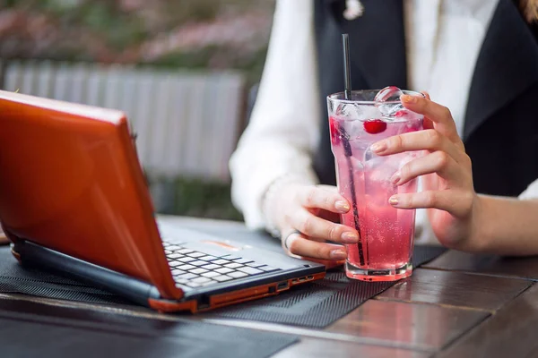 Una joven estudiante bebe limonada fresca mientras trabaja remotamente en una computadora portátil en un café en un soleado parque de la ciudad — Foto de Stock