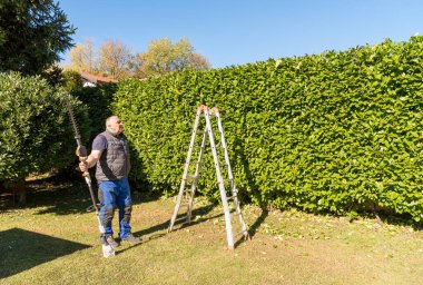 Bir ev bahçesinde yeşil bir çitin önünde elektrikli çit biçme makinesi tutan olgun bir adam..