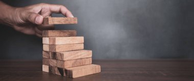 Yakın plan el, çöküşü veya finansal risk yönetimi ve stratejik planlama kavramlarını önlemek için piramit şeklinde dizilmiş ahşap blok kulesini ihtiyatlı bir şekilde yerleştiriyor.