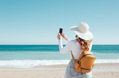 Kumsalda akıllı telefonunu, şapkasını ve sarı sırt çantasını kullanan bir kadın.