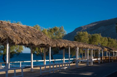 Santorini 'deki kara kum plajı. Kapatma sırasında. Boş sokak. Kıyıda yeşil ağaçlar. Kocaman bir kaya. Hiç kimse. Saman şemsiyeler.