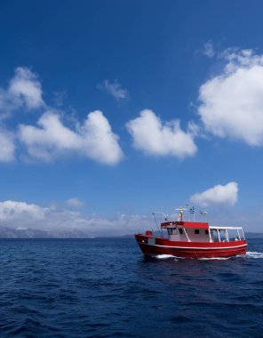 Mavi deniz suyunda küçük kırmızı bir balıkçı teknesi. Arkaplanda adaların siluetleri.