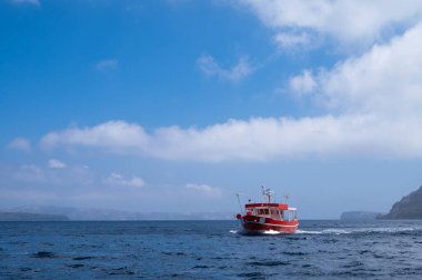Mavi deniz suyunda küçük kırmızı bir balıkçı teknesi. Arkaplanda adaların siluetleri.
