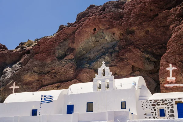 White church with bell tower and blue doors at the foot of red rock. Bottom view.Traditional Orthdox church on Santorini island, Greece.