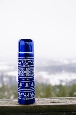 Ahşap yüzeyinde desenleri olan mavi termos şişesinin yakın çekimi. Arka planda bulanık kış ormanı. Açık hava pikniği.