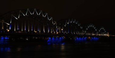 Riga, Letonya 'daki Daugava nehri üzerindeki aydınlık demiryolu köprüsü. Gece şehir manzarası.