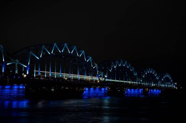 Riga, Letonya 'daki Daugava nehri üzerindeki aydınlık demiryolu köprüsü. Gece şehir manzarası.