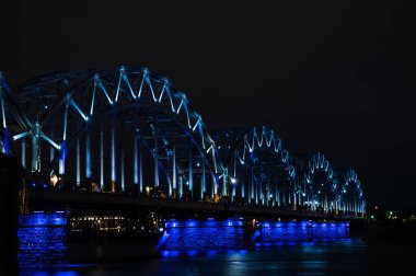 Riga, Letonya 'daki Daugava nehri üzerindeki aydınlık demiryolu köprüsü. Gece şehir manzarası.