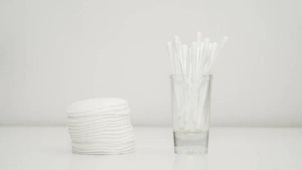 Close-up of stack of cotton pads and cotton swabs in a glass cup. White background.