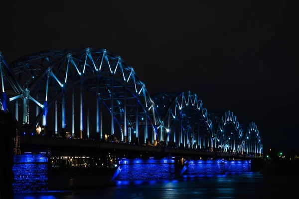 Riga, Letonya 'daki Daugava nehri üzerindeki aydınlık demiryolu köprüsü. Gece şehir manzarası.