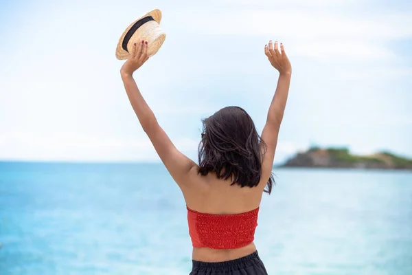 Back woman tan skin wearing pink tank top hold a straw hat with standing arms outstretched on sky. looking into the sea and fresh sky. Summer travel. Relax, Holiday and tropical, comfortable concept.