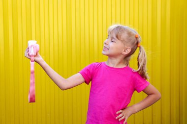 Pembe tişörtlü şirin küçük kız pembe bir kamerada selfie çekiyor ve gülümsüyor. Kopyalama alanı olan sarı arkaplan. Küçük bir kızın duyguları. Bir çocuk fotoğrafçılığın temellerini öğrenir..