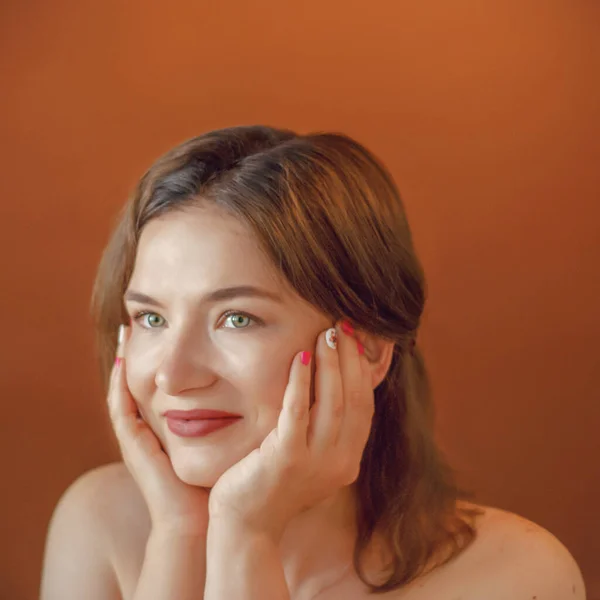 large portrait of a European girl with hands on a brown background. One girl smiles on a plain background. 30 years old. Face. Hand.look, eyes. Face on your hands.