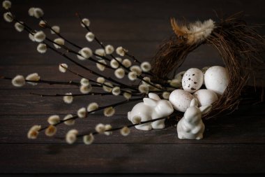 Fotokopi alanı olan mutlu Paskalyalar. Yuvada dekoratif yumurtalar, söğüt dalları ve koyu kahverengi ahşap arka planda seramik tavşan figürleri olan şenlikli bir kompozisyon..