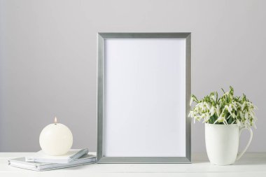 Hello bahar kartı modeli ya da modern İskandinav tarzında seramik bardakta buket kardelen çiçekleri ve ahşap masada yanan mumlarla dolu gümüş fotoğraf çerçevesi. Kopyalama alanı olan beyaz arkaplan