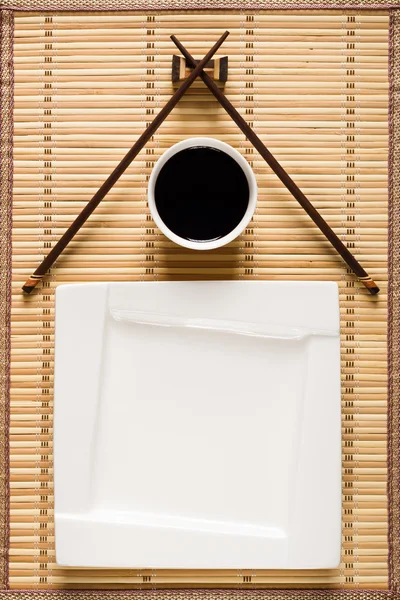 Палочки, белая тарелка и миска с соевым соусом на бамбуковом коврике — стоковое фото