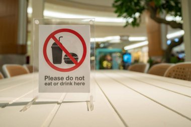 Kalabalık bir açık alanda, beyaz bir masada, ziyaretçilere yemek ve içki içmemeleri gerektiğini hatırlatan açık bir güvenlik işareti. Tabela çerçevelenmiş ve kırmızı bir yasak içeriyor.