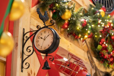 Ahşap bir duvara monte edilmiş, içinde horoz rüzgar gülü ve asılı bir çan olan büyüleyici bir antika saat. Etrafı şenlikli Noel süslemeleri ve kırsal dekorlarla çevrili ve rahat bir tatil yaratıyor..