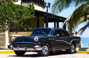 HAVANA,CUBA - JUNE 23, 2014: Cuba black american vintage car parked under palms and before a front of a building clipart