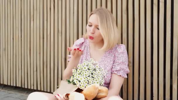 Elegante donna bionda con capelli corti e rossetto rosso rilassante all'aria aperta. Ragazza carina con camomilla mazzo e sacchetto di carta baguette francese. Allegramente strappando petali e soffiando, guardando la fotocamera — Video Stock