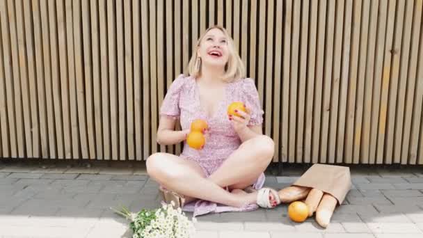 Zabawna kobieta żonglująca pomarańczami w plenerze, siedząca na podłodze ze skrzyżowanymi nogami. Happy girl wygłupiając się i zabawy rzucanie owoców rąk na tle drewniane ściany, kilka rumianku i bagietka torba papierowa — Wideo stockowe