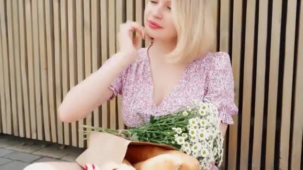 Молодая привлекательная блондинка чинит волосы в солнечный день на улице. Девушка в летнем платье расслабляется и улыбается сидя на открытом воздухе, держа с кучей ромашки и французский багет в бумажном пакете — стоковое видео
