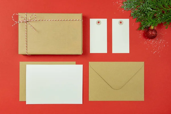 Concepto de carta navideña. Tarjeta de papel en blanco, sobre de papel artesanal, etiqueta, caja de regalo vintage, cuerda, ramas de thuja sobre fondo rojo. Vista superior. Copiar espacio. Burla.. — Foto de Stock