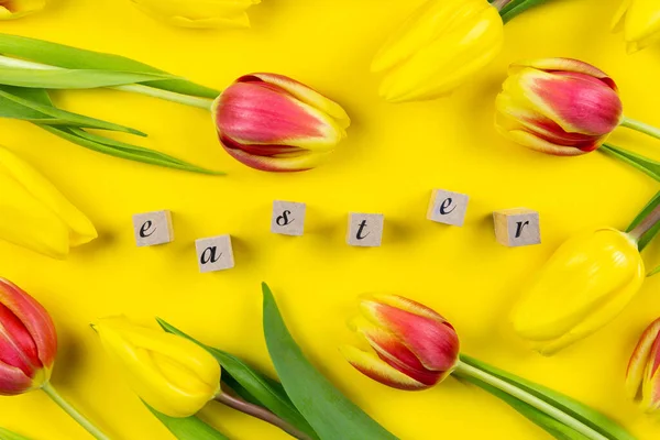 Easter floral background with colorful tulips and word easter using wooden blocks on yellow background. Top view.