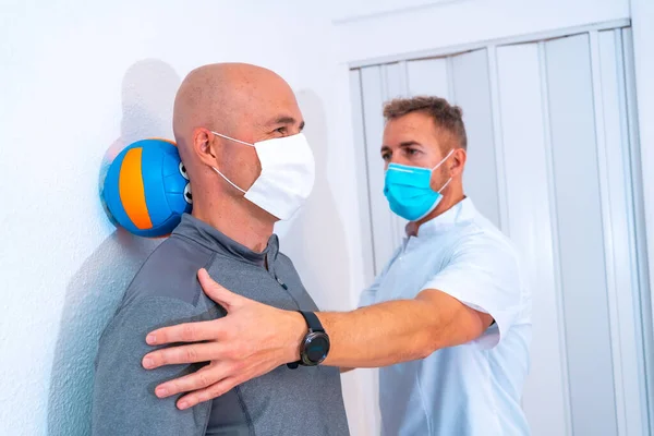 Maskeli fizyoterapist, müşterinin boynundaki kılları uzatmasına yardım ediyor. Koronavirüs salgınında koruyucu önlemlerle fizik tedavi, COVID-19. Osteopati, spor kiromasaj