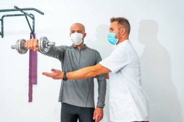 Yüz maskeli bir fizyoterapist ve halterle egzersiz yapan bir hasta. Koronavirüs salgını için koruyucu önlemlerle fizik tedavi, COVID-19. Osteopati, spor kiromasaj