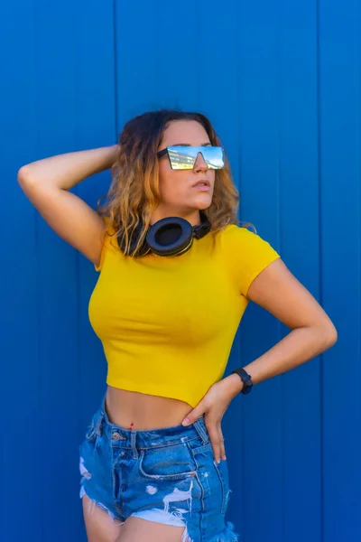 生活方式金发白人女孩穿着蓝色背景的黄色T恤戴着音乐耳机和太阳镜的年轻女子 — 图库照片
