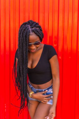 Yaşam tarzı, siyah tişörtlü siyah kız, kot şort ve kırmızı arka planda güneş gözlüğü. Genç ve şehvetli bir kapan dansçısının portresi.