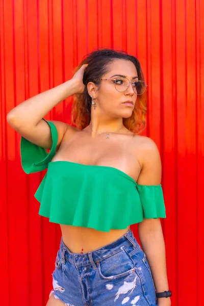 Lifestyle, Caucasian blonde girl in green t-shirts, denim shorts and transparent see glasses on a red background. With a seductive look at the summer photoshoot