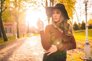 Autumn lifestyle at sunset, blonde Caucasian woman in a red sweater and black hat, enjoying nature in a park with trees. On a lovely city park trail clipart