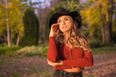 Lifestyle, blonde Caucasian girl in a red sweater and black hat, enjoying nature in a park with trees, portrait of the young woman enjoying the autumn sun clipart