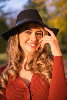 Autumn lifestyle, blonde Caucasian girl in red sweater and gorgeous black American hat, enjoying nature in park with big trees, portrait of young model smiling clipart