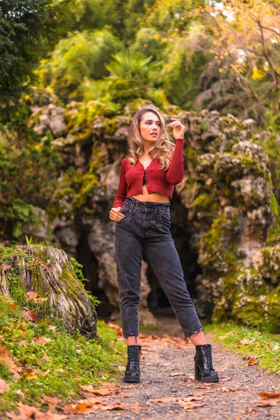 秋天的生活方式 穿着红色毛衣 牛仔裤 高跟鞋的金发白人女孩 在一个有着自然洞穴的公园里欣赏大自然 — 图库照片