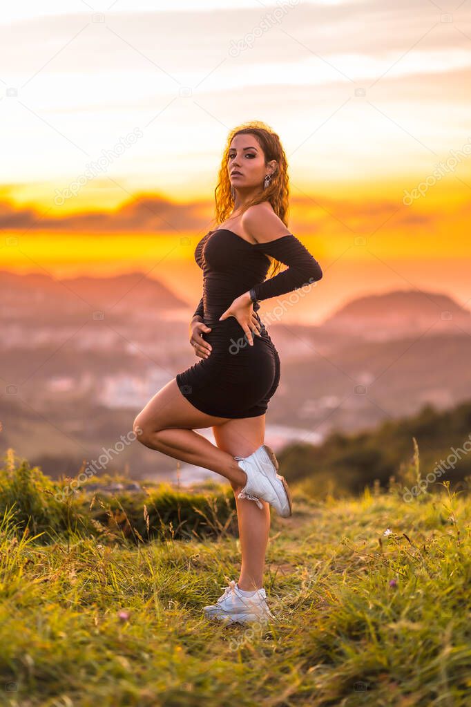Summer lifestyle, a young Caucasian woman with wavy brown hair in a tight black dress, the young woman posed in the sunset on top of a mountain