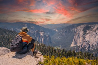 A young man on the trek sitting on the Sentinel Dome lookout in Yosemite National Park looking at El Capitan Mountain. United States clipart