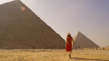 Kırmızı elbiseli ve hasır şapkalı genç bir turistin Giza piramitlerinde yürürken görüntüsü. Kahire, Mısır
