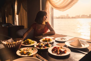 Genç bir turist, Nil 'de bir teknede yemek yiyor. Penceresinde günbatımı ışığı olan geleneksel bir Mısır yemeği. Afrika